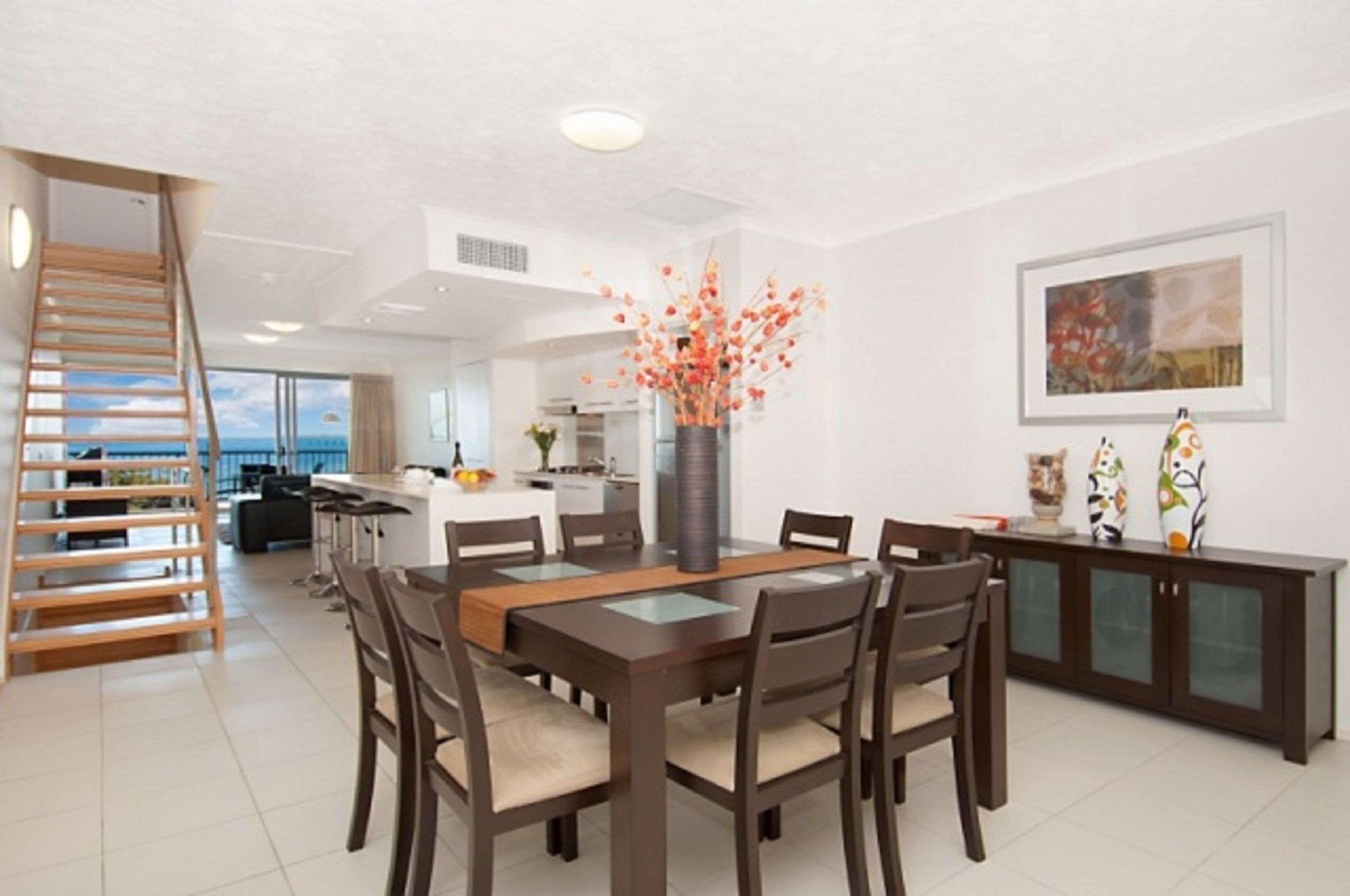 A modern dining area with a wooden table and chairs, a vase with orange flowers, and a view of the ocean through large windows.