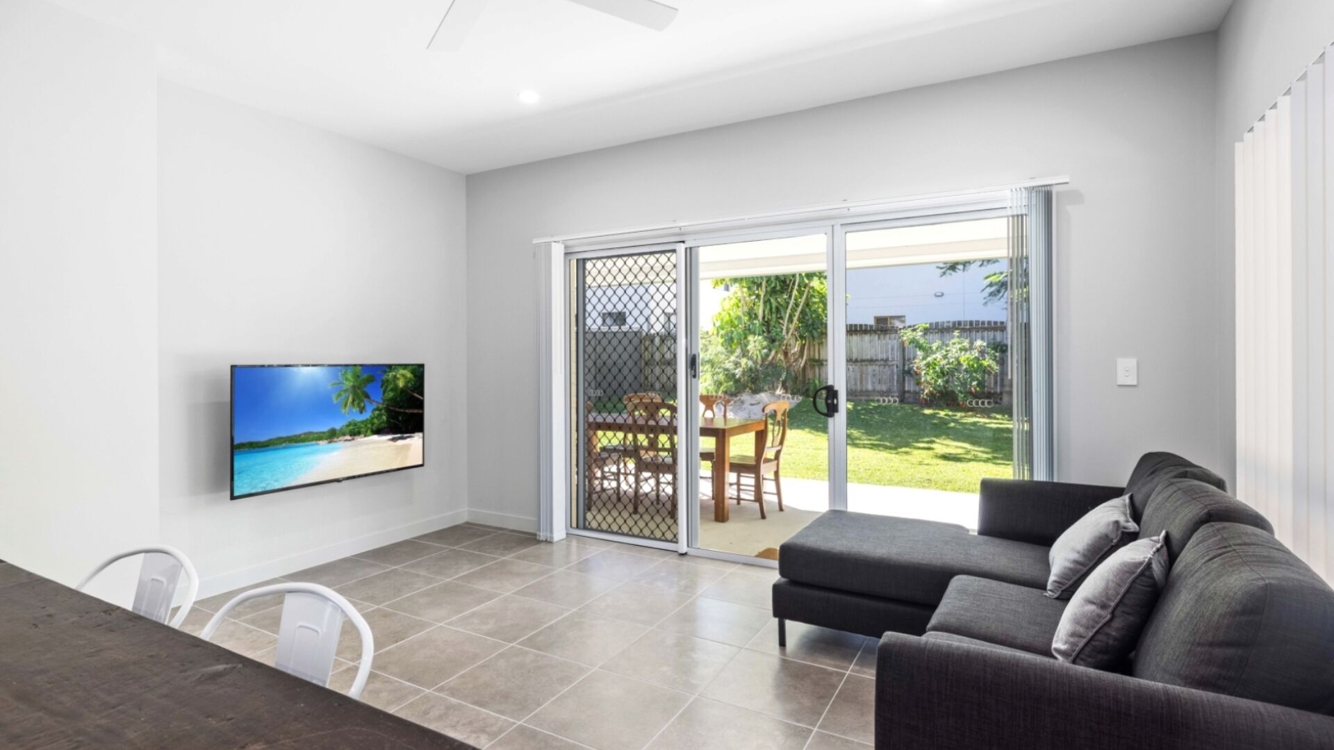 A modern living room with a flat-screen TV, a dark sofa, and a sliding glass door leading to a patio with outdoor furniture.