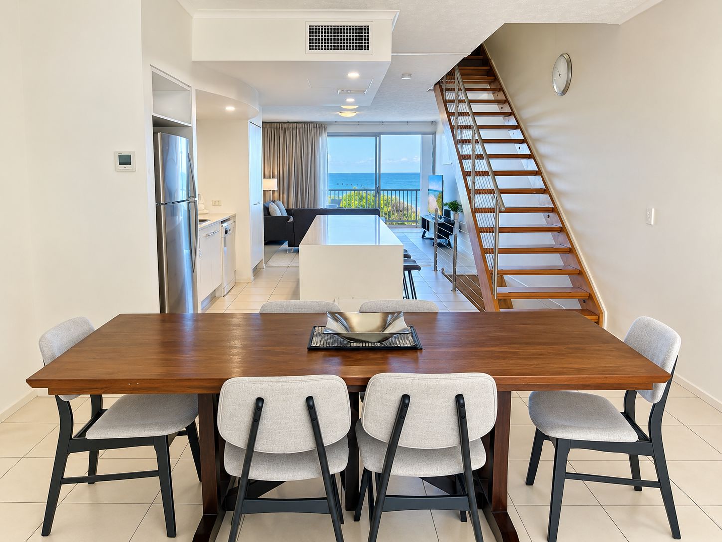 Uma sala de jantar moderna com mesa de madeira e quatro cadeiras, cozinha com geladeira, e escada que leva a uma varanda com vista.