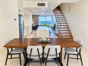 Un espace repas moderne avec une table en bois et quatre chaises, une cuisine équipée d’un réfrigérateur, et un escalier menant à un balcon avec vue.