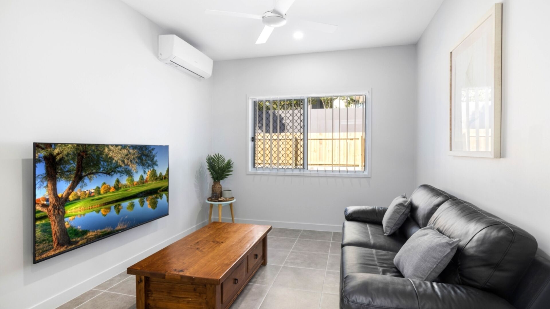 A living room with a wooden coffee table, a black leather sofa, a mounted TV, a ceiling fan, and a window with a view of a fence.