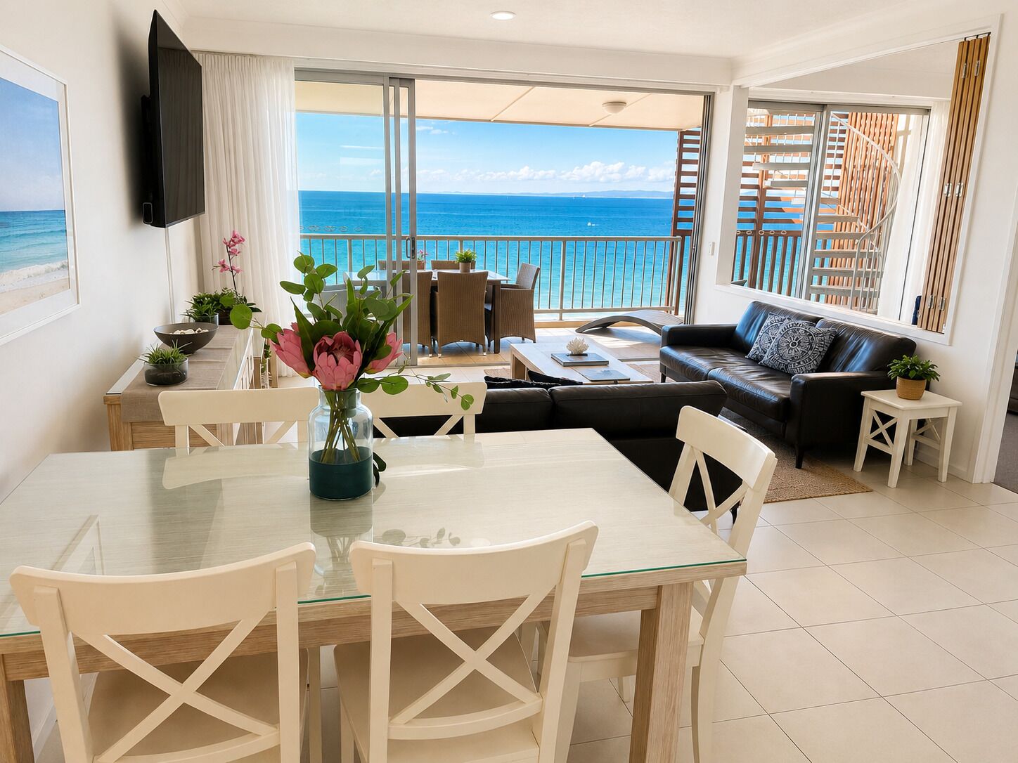 Ein modernes Wohnzimmer mit Balkon und Meerblick, eine Essecke mit Glastisch sowie eine Couch mit Aussicht.
