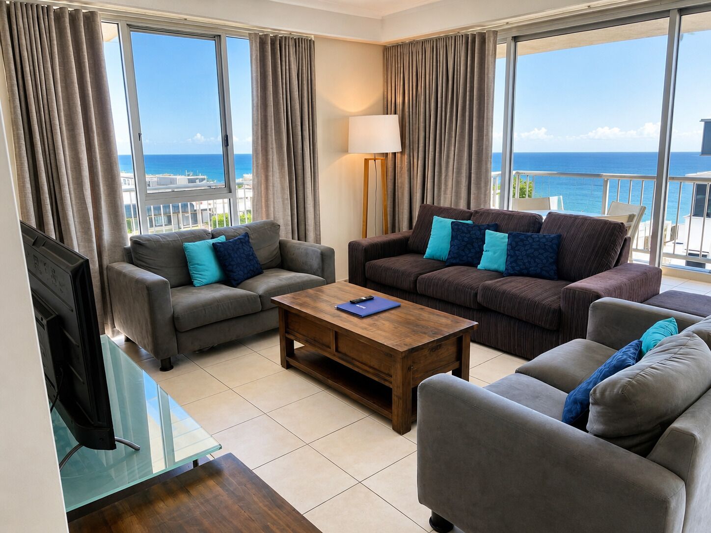 Ein modernes Wohnzimmer mit einer Sofagarnitur, einem Couchtisch und einem Fernseher. Große Fenster bieten Blick auf den Ozean.