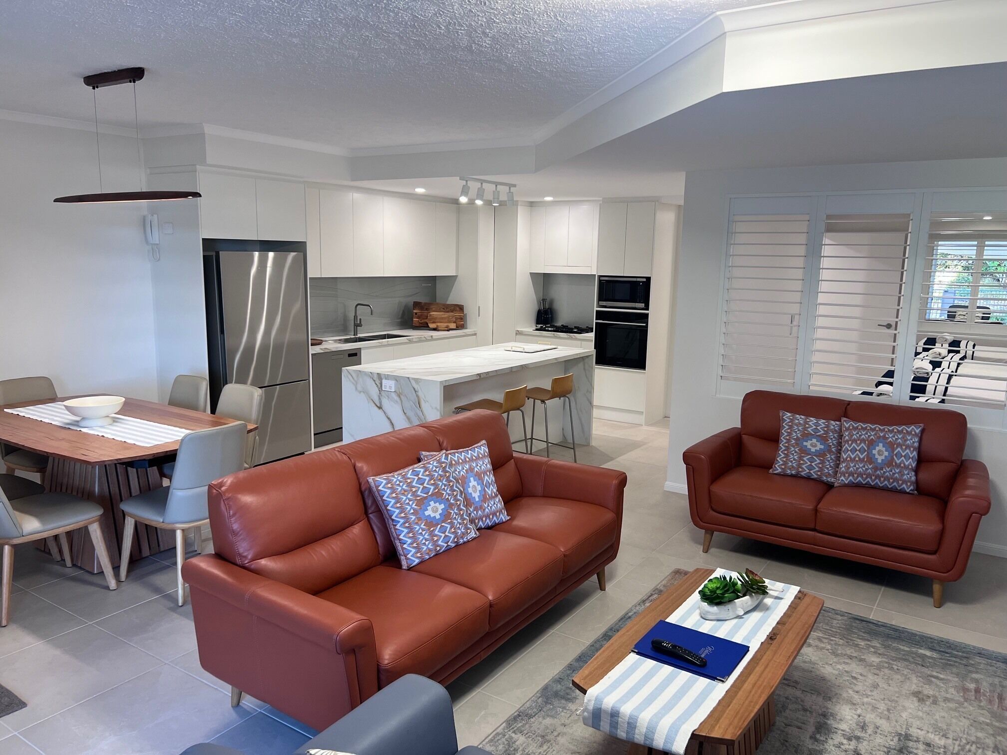 A modern living room with a brown leather sofa, a coffee table, and a dining area with a white table and chairs.