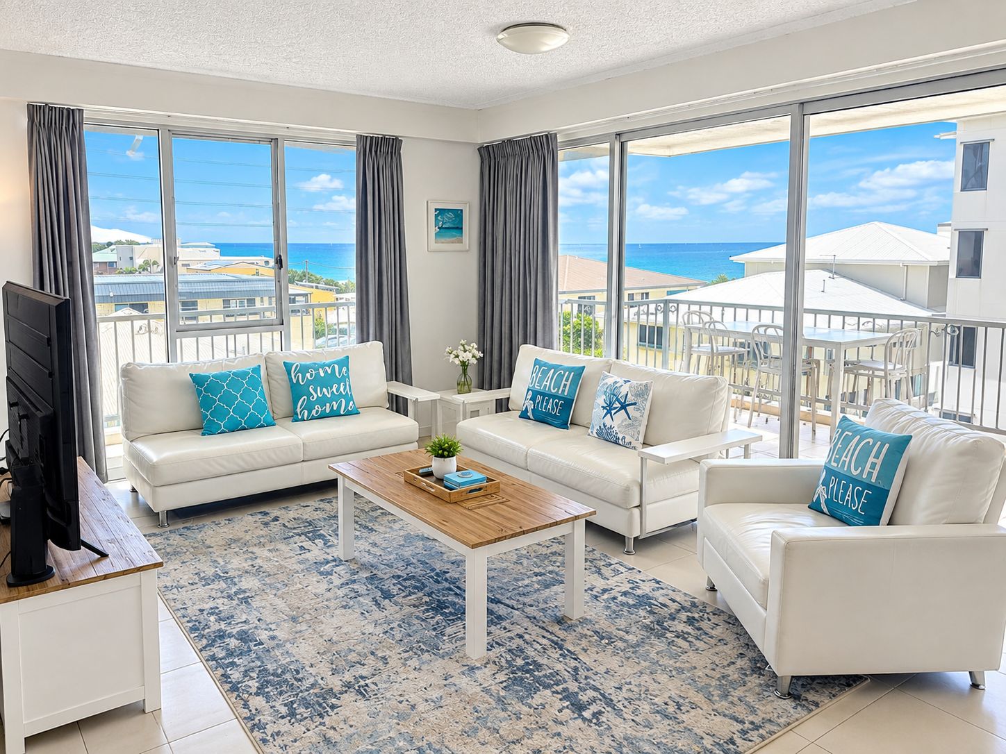 A spacious living room with a large window overlooking the sea, two white sofas with blue pillows, a wooden coffee table, and a TV on the left.