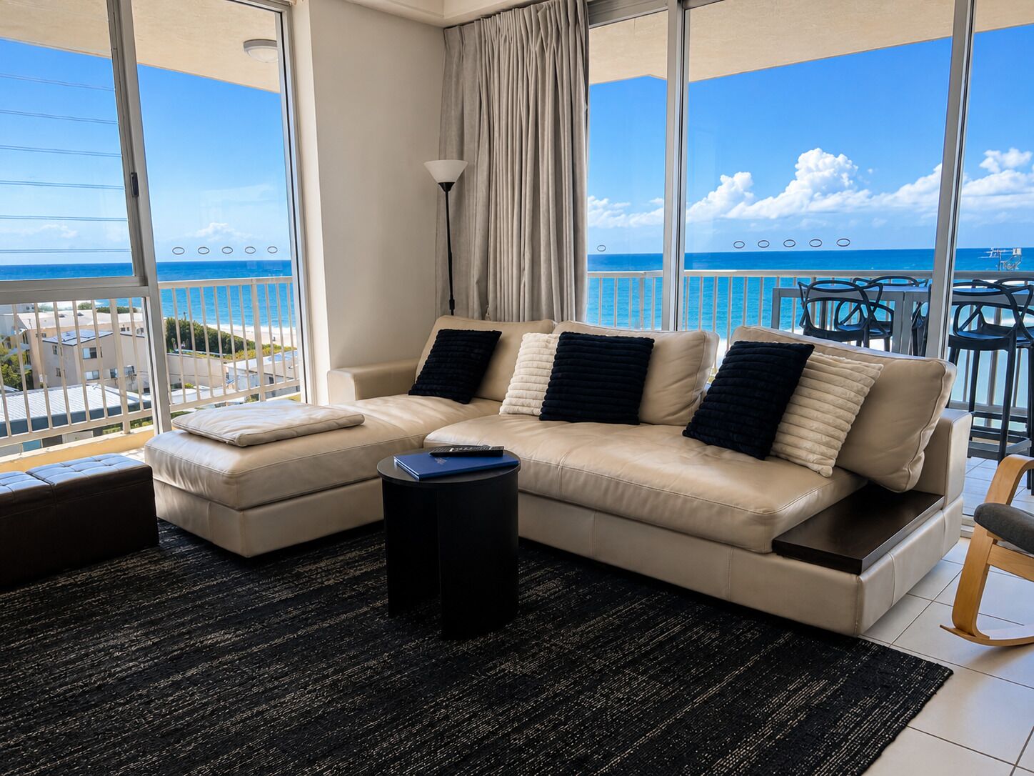 A modern living room with a beige sofa, black cushions, a black coffee table, and a view of the ocean.