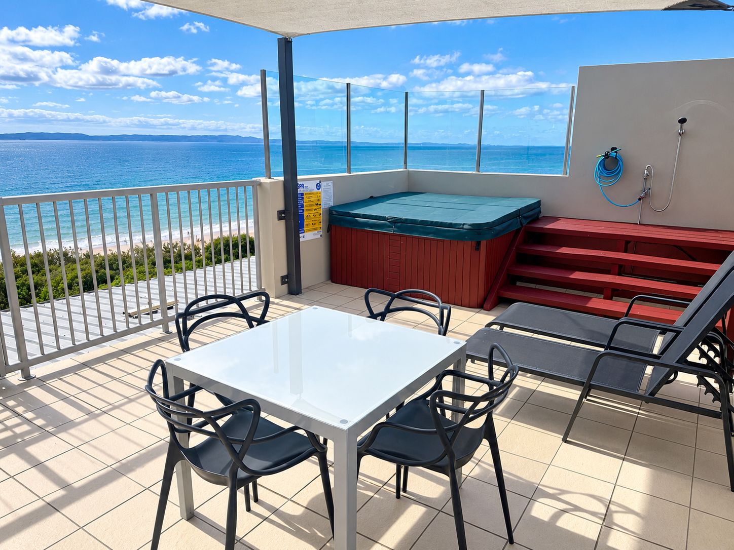 A balcony with a table and chairs, a hot tub, and a view of the beach.
