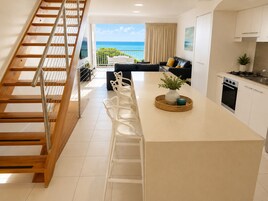Una cocina moderna con una isla central, un área de comedor con sillas blancas, una escalera y vistas al océano.