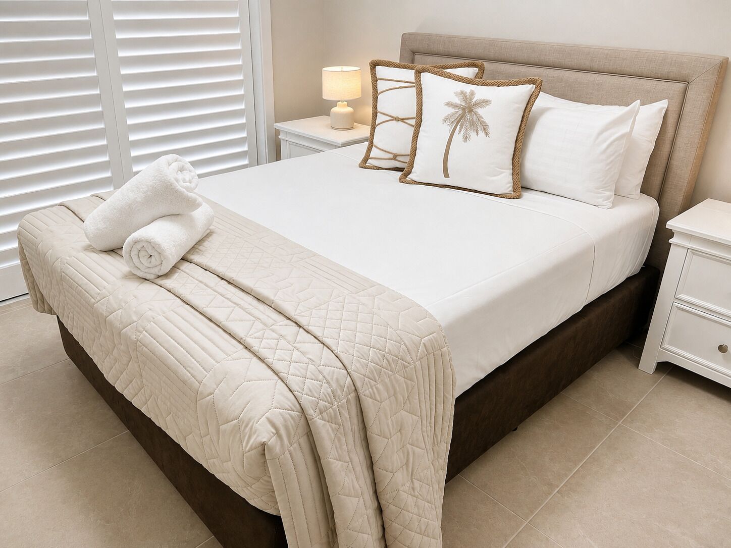 A neatly made bed with a quilted cover, a rolled towel, and two pillows with a palm tree design.