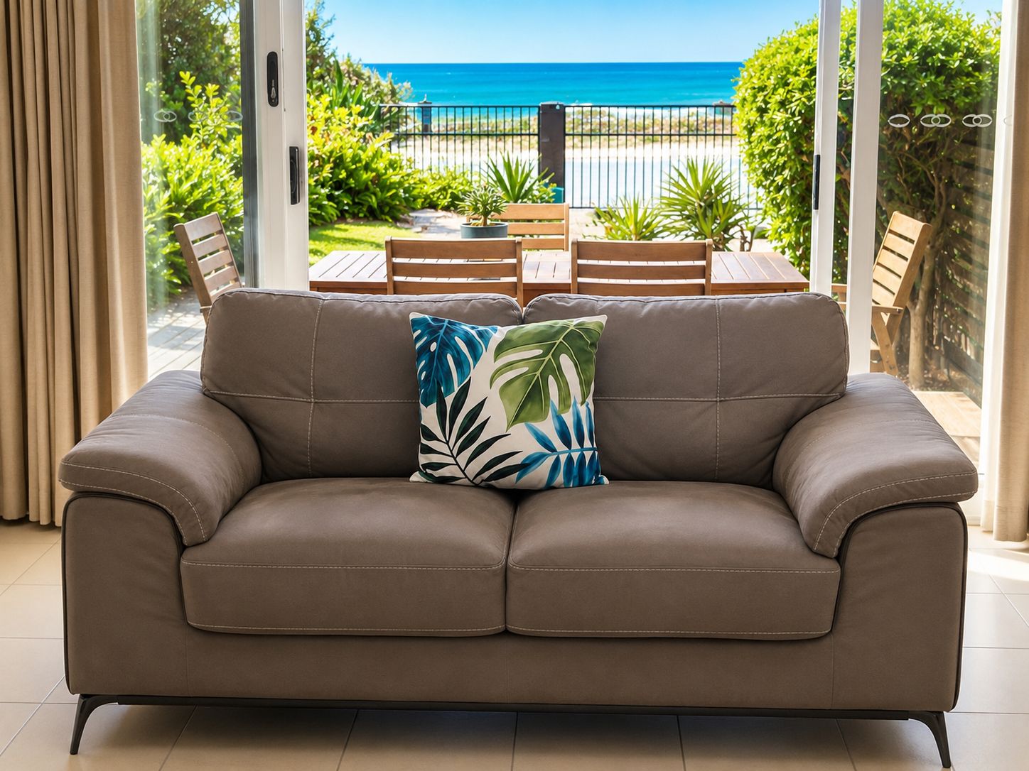 Um sofá de couro marrom com almofada em formato de folha tropical, em um quarto com vista para a praia e vegetação.