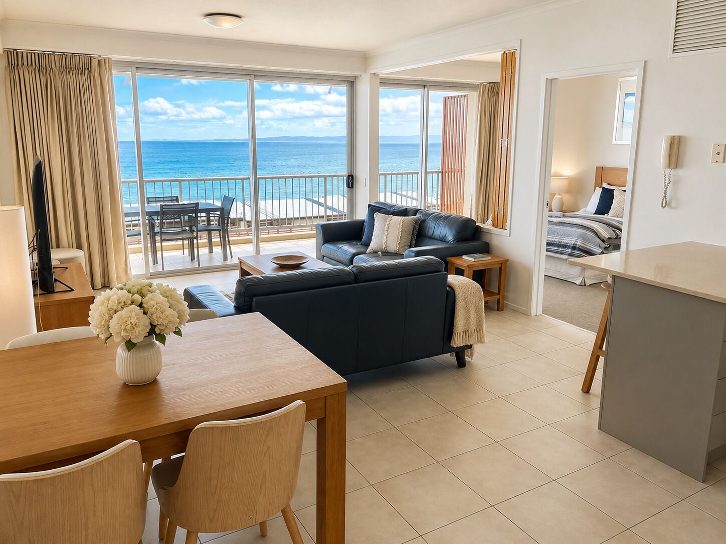 Un salon moderne avec un canapé, une table à manger et des chaises, un balcon offrant une vue sur la mer, et une chambre à coucher avec un lit et une table de chevet.