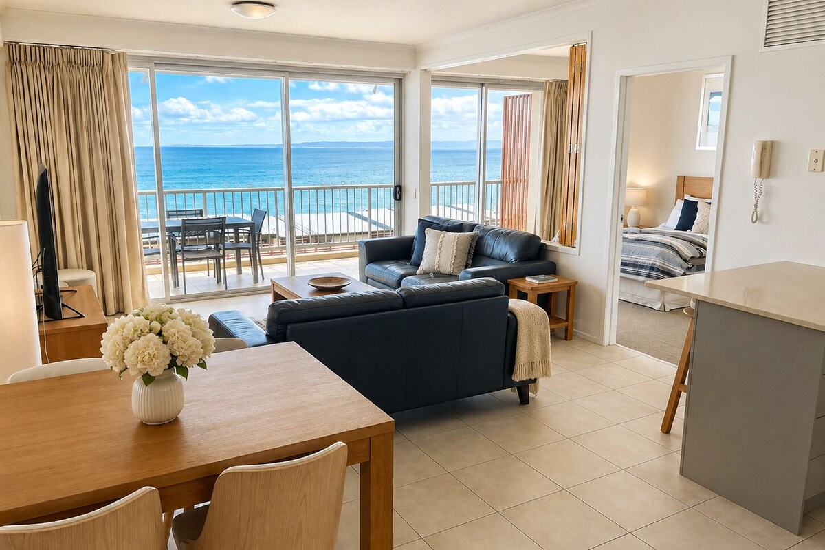 Un salon moderne avec un canapé, une table à manger et des chaises, un balcon offrant une vue sur la mer, et une chambre à coucher avec un lit et une table de chevet.