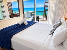 Habitación de hotel con cama, vista al océano, balcón con mesa y sillas, y ventana con cortinas.