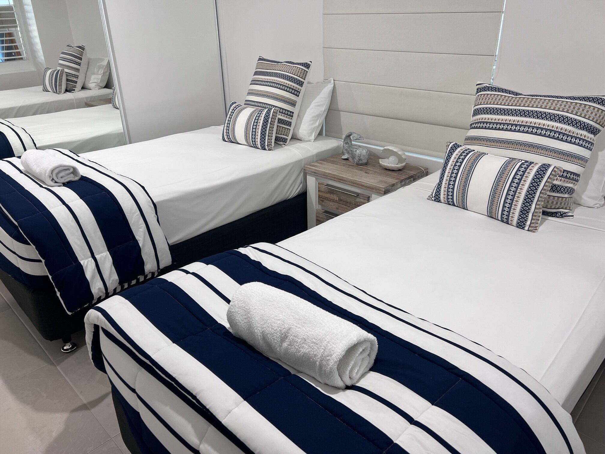 A hotel room with two beds, each with striped bedding and pillows, a bedside table with a lamp, and a mirror on the wall.