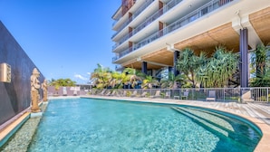 A clear pool with a curved edge, surrounded by lounge chairs and palm trees, with a multi-story building in the background. - On the Beach Resort Bribie Island (Woorim)