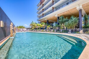 Una piscina cristalina de borde curvo, rodeada de sillas de descanso y palmeras, con un edificio de varios pisos al fondo.