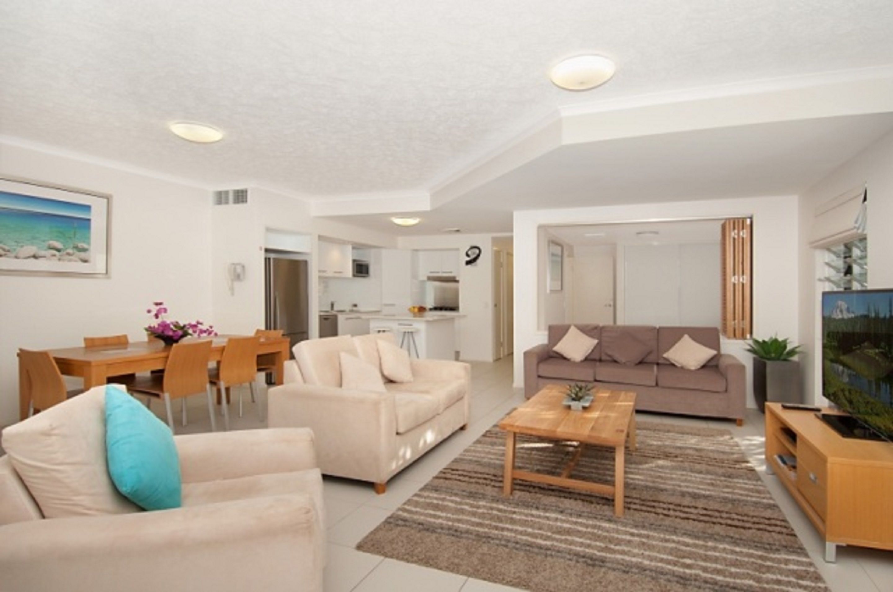 A modern living room with a sofa, coffee table, and TV stand. There is a dining area with a table and chairs, and a kitchen in the background.