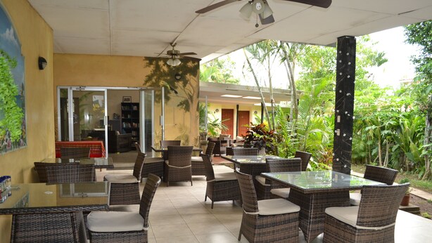 Un patio al aire libre con mobiliario de mimbre y mesas de vidrio, rodeado de abundante vegetación y vistas del interior.