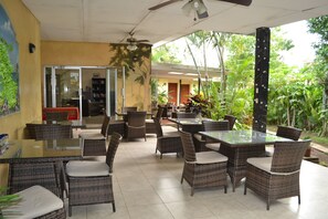 Un patio al aire libre con mobiliario de mimbre y mesas de vidrio, rodeado de abundante vegetación y vistas del interior.