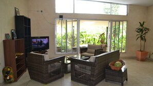 Una sala de estar con mobiliario de mimbre, televisor y puerta corrediza de vidrio que da a un balcón con plantas.