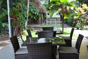 Uma mesa com tampo de vidro e cadeiras de vime em ambiente externo, com vegetação e estrutura de telhado de palha.