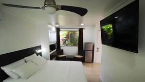 Una habitación de hotel con cama, ventilador de techo, televisor, refrigerador y ventana con cortinas.