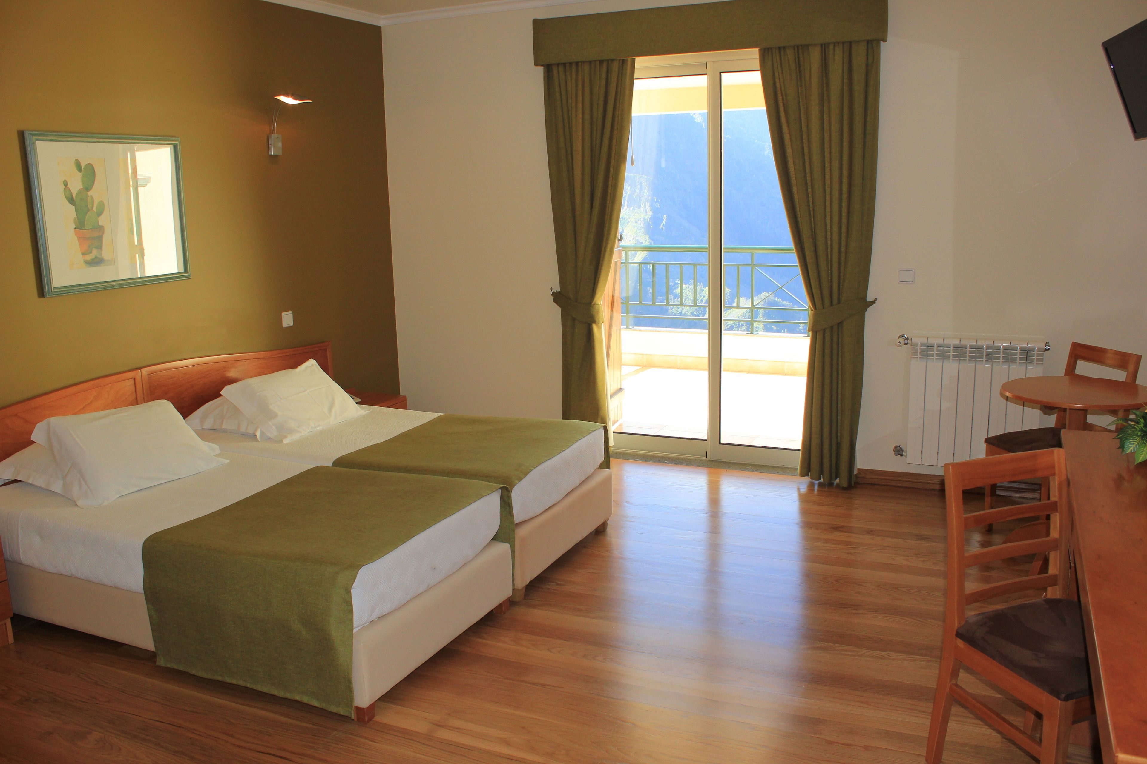 Quarto de hotel com cama de casal, uma pequena mesa com cadeira, varanda e vista, e radiador.