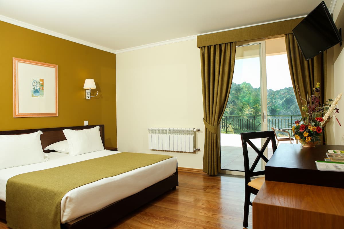 Hotelkamer met een bed, bureau, stoel, televisie, raam met gordijnen, radiator en een lijst aan de muur.