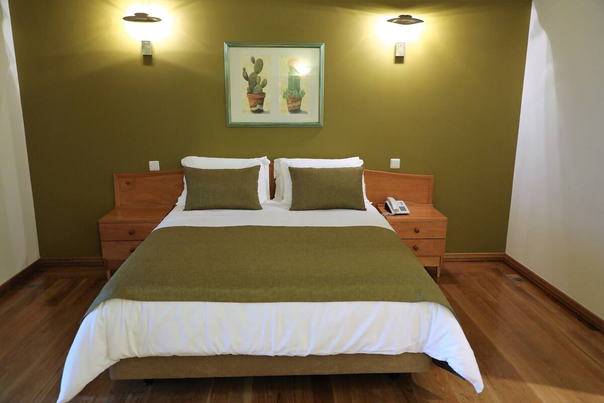 Een netjes opgemaakt bed met een groene deken, twee nachtkastjes met lampen, en een lijst met foto’s van cactussen aan de muur.