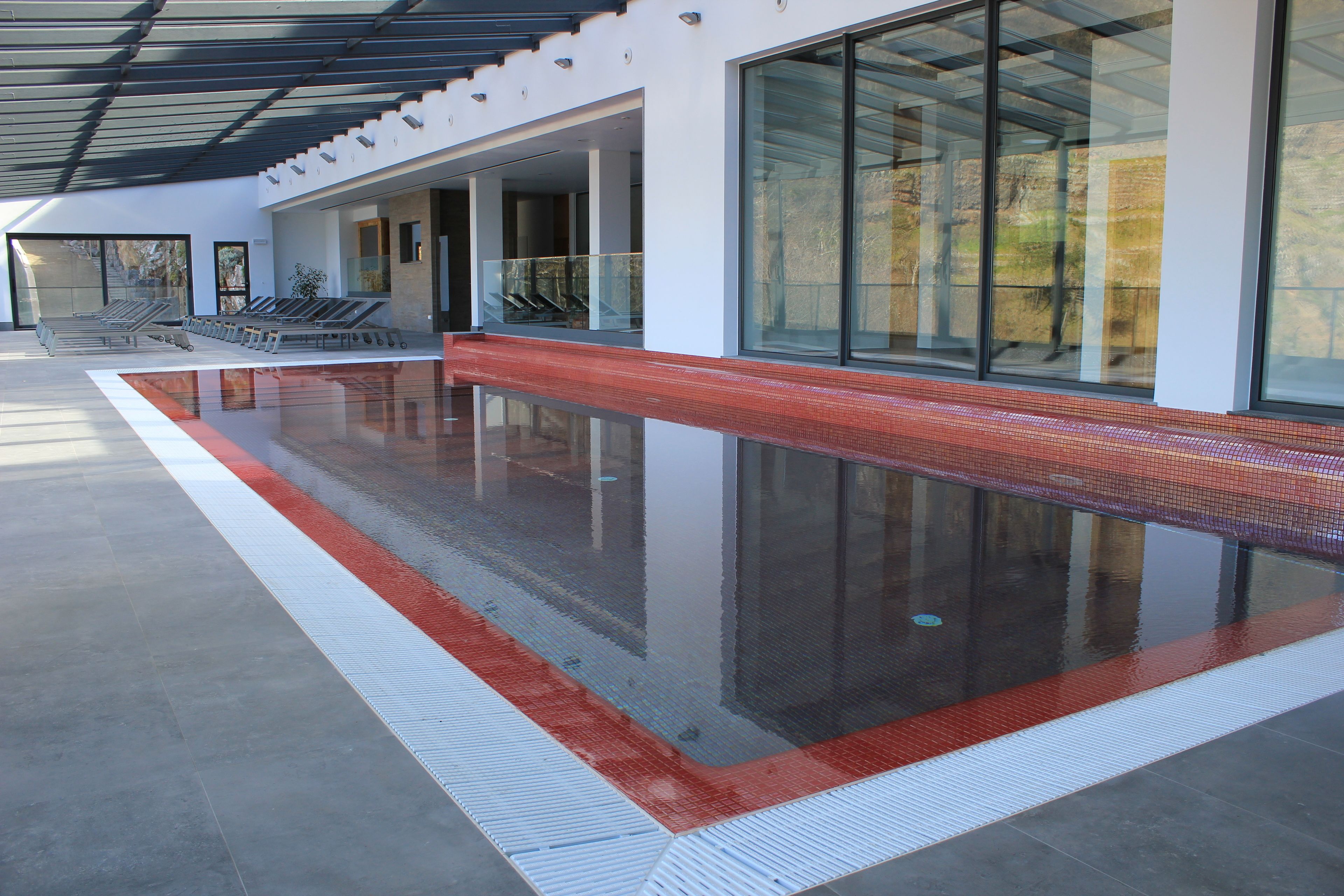 Une piscine intérieure moderne avec des parois en verre transparent, un bord carrelé en rouge et blanc, et un sol en béton.