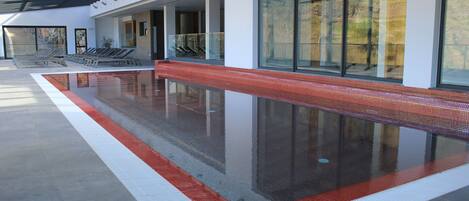 Une piscine intérieure moderne avec des parois en verre transparent, un bord carrelé en rouge et blanc, et un sol en béton.