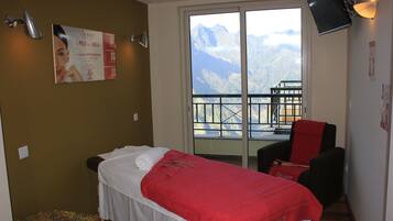 A bedroom with a bed, a red blanket, a massage table, a chair, and a balcony with a mountain view.