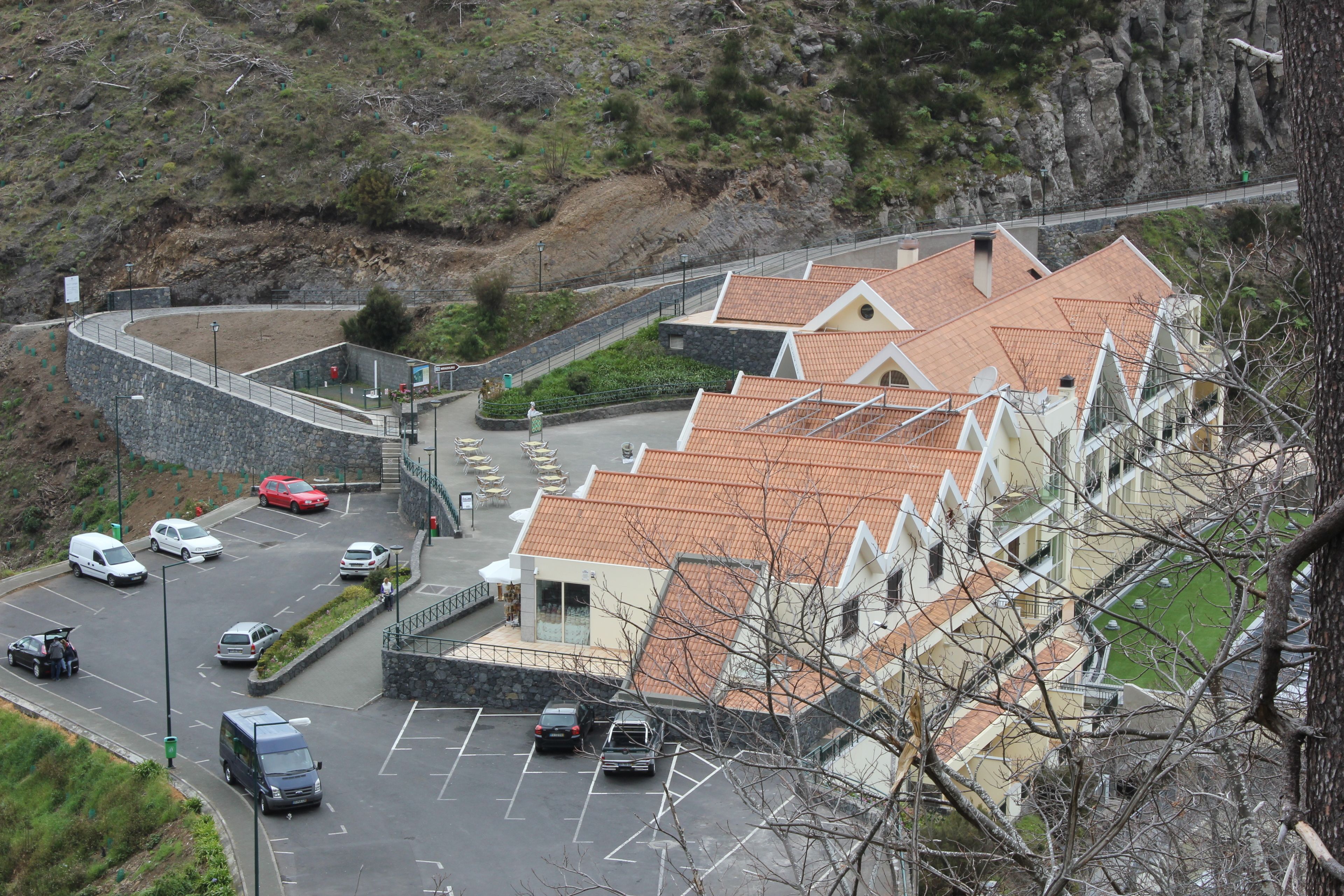 Un estacionamiento con varios coches, un edificio con techo de tejas y un muro de contención de piedra.