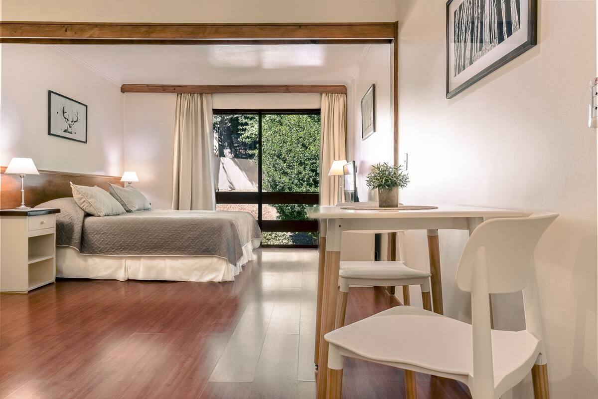 A bedroom with a bed, bedside tables, a desk, and a chair. There is a window with curtains and a view of greenery outside.