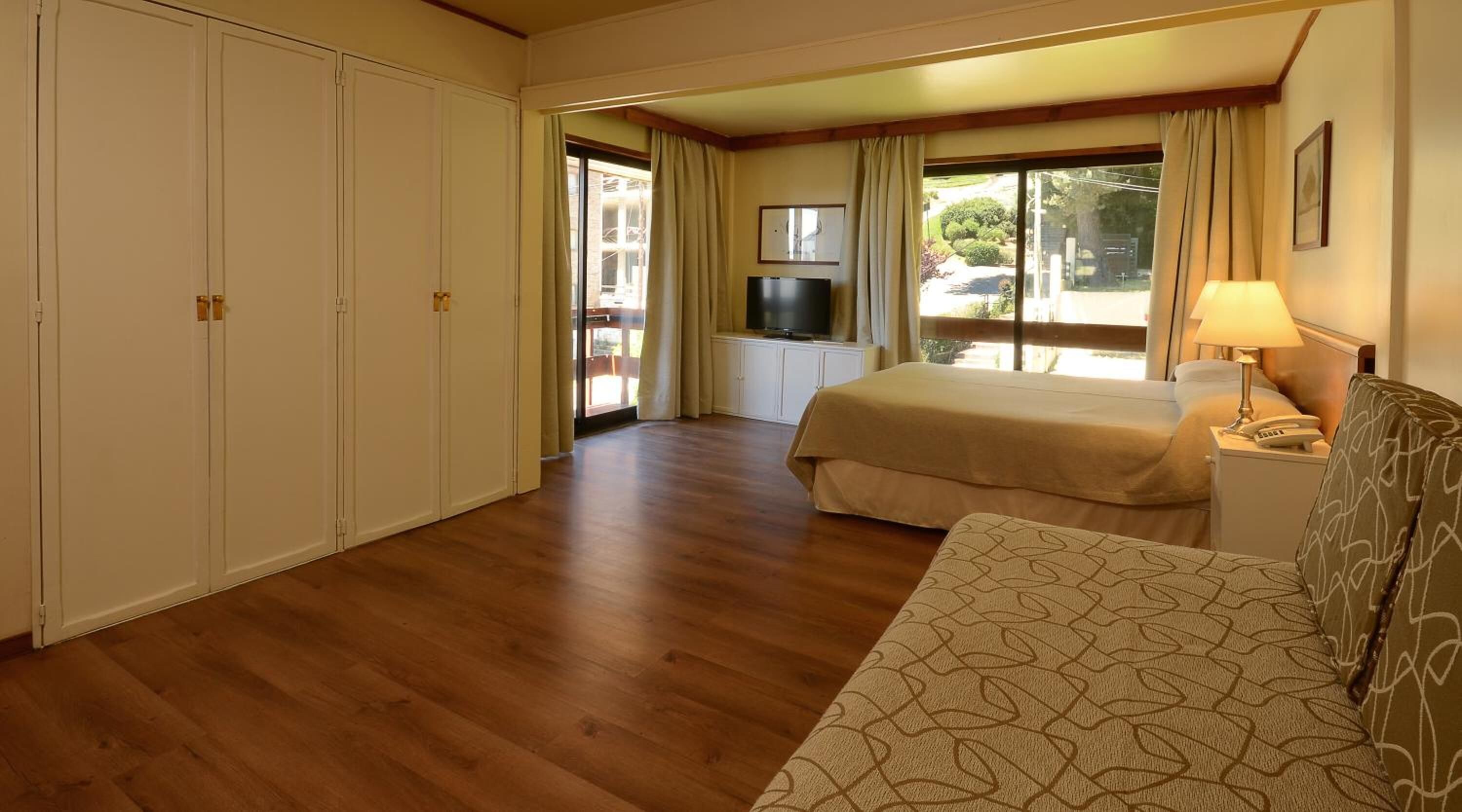 A hotel room with a wooden floor, a bed with a beige bedspread, a sofa with a patterned cushion, a TV on a cabinet, and a window with curtains.