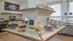 Un mostrador de buffet con diversos postres y pasteles, un letrero en una pizarra y una zona de cocina al fondo.