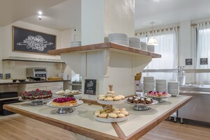 Un mostrador de buffet con diversos postres y pasteles, un letrero en una pizarra y una zona de cocina al fondo.