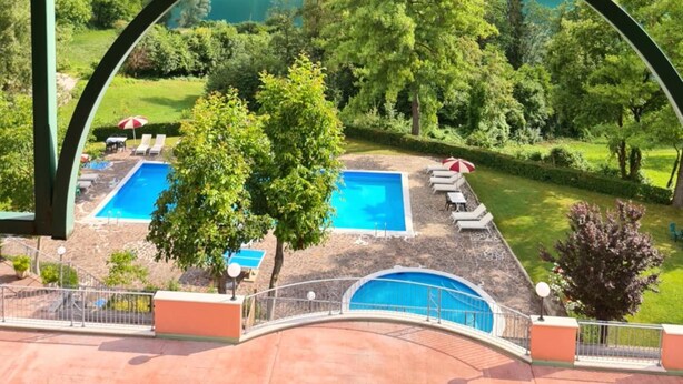 Vista attraverso un'ampia finestra su un paesaggio montano con due piscine e lettini.