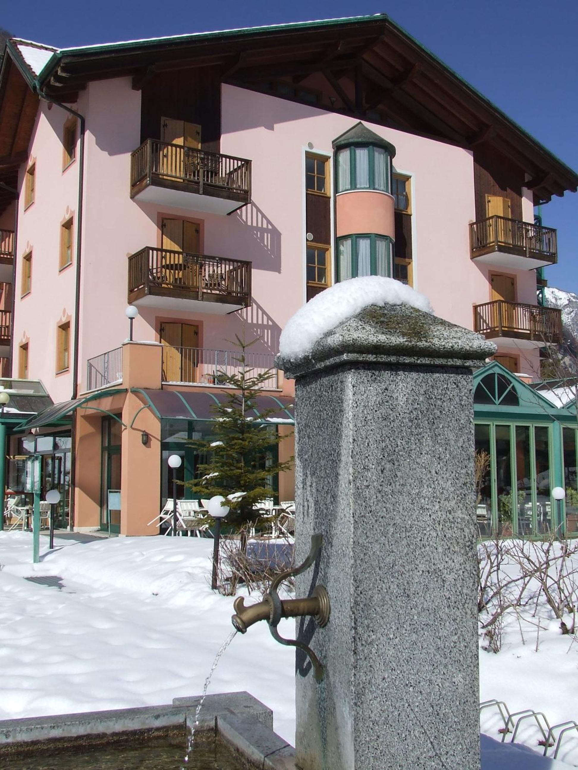 Un caño de piedra con un pomo metálico, suelo cubierto de nieve y un edificio con balcones y techo verde.