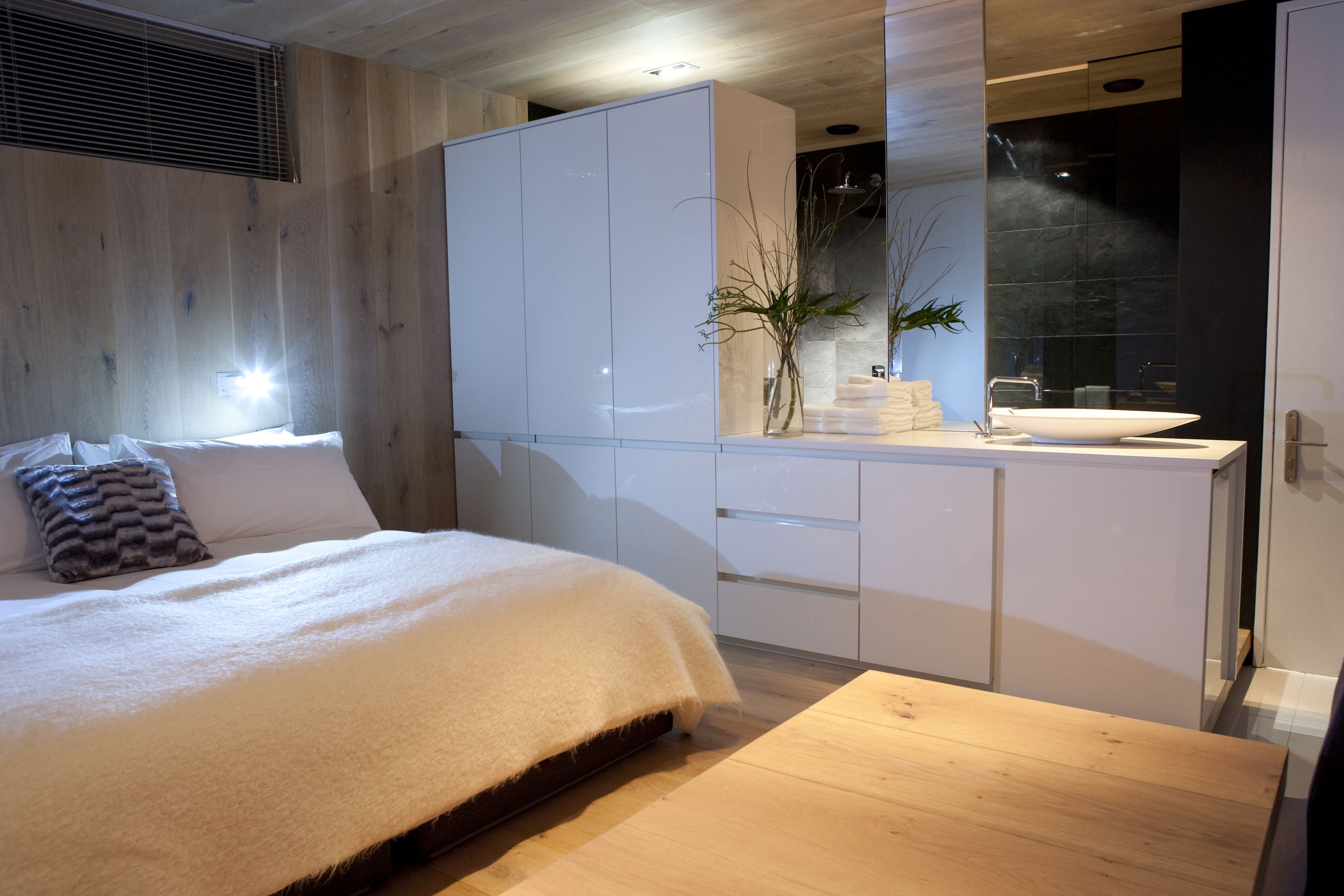 Une chambre moderne avec un grand lit, un plancher en bois, une armoire blanche, une salle de bain dotée d’un grand miroir et d’un lavabo, et une petite plante sur la commode.