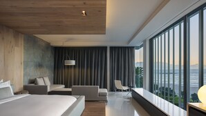 Et moderne hotelværelse med en stor seng, et siddeområde med sofa og stole, udsigt over havet og et træloft.