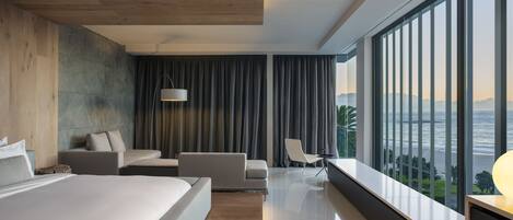 A modern hotel room with a large bed, a seating area with a sofa and chairs, a view of the ocean, and a wooden ceiling.