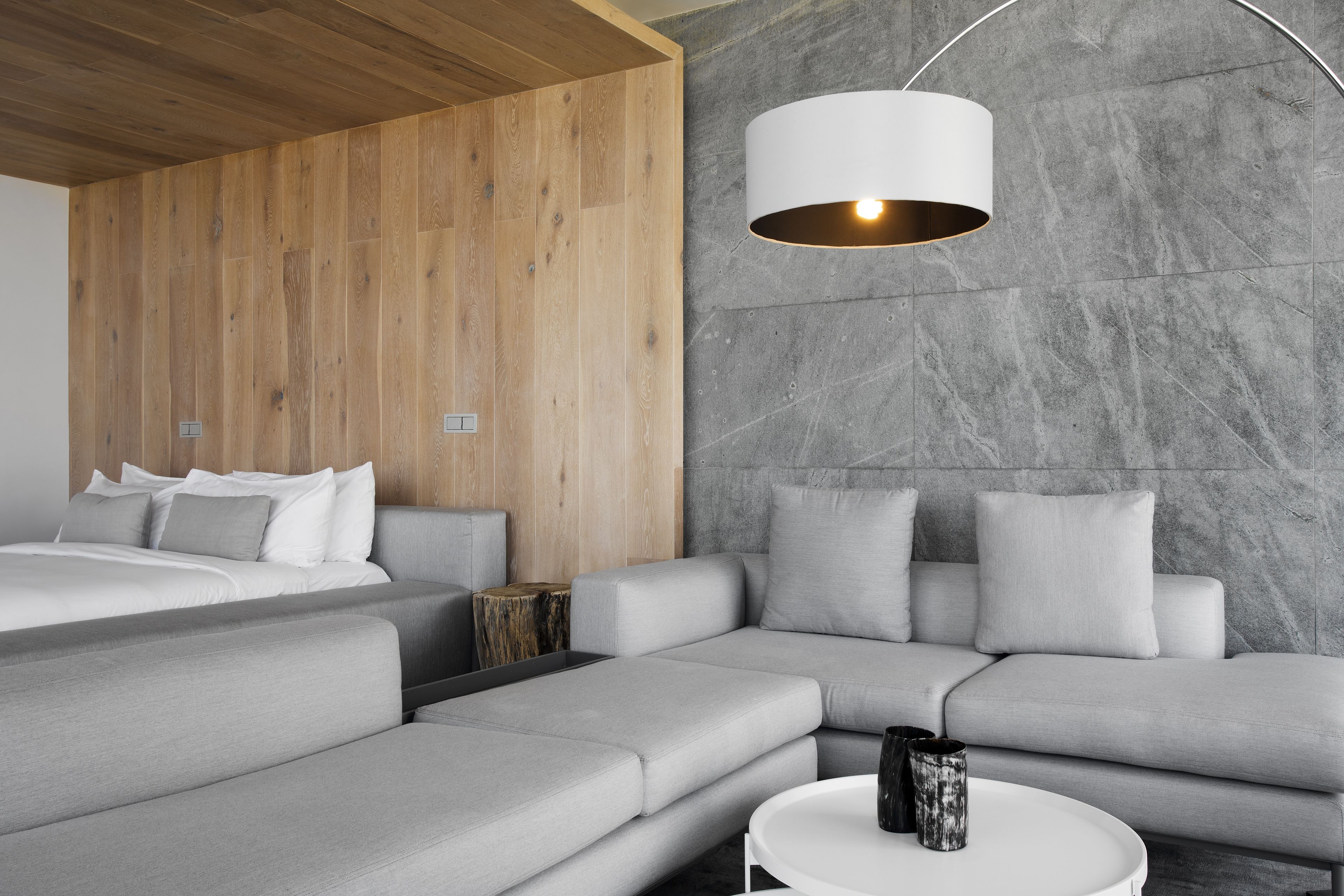 Una habitación de hotel moderna con techo de madera, sofás grises, una cama con ropa de cama blanca y una pared de mármol.