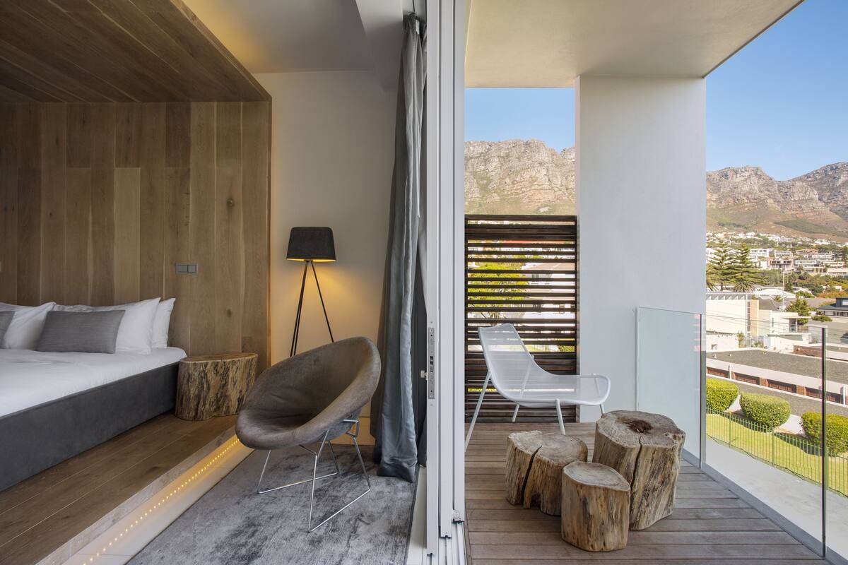A modern bedroom with a large bed, a chair, a small table, and a balcony with a view of the city and mountains.
