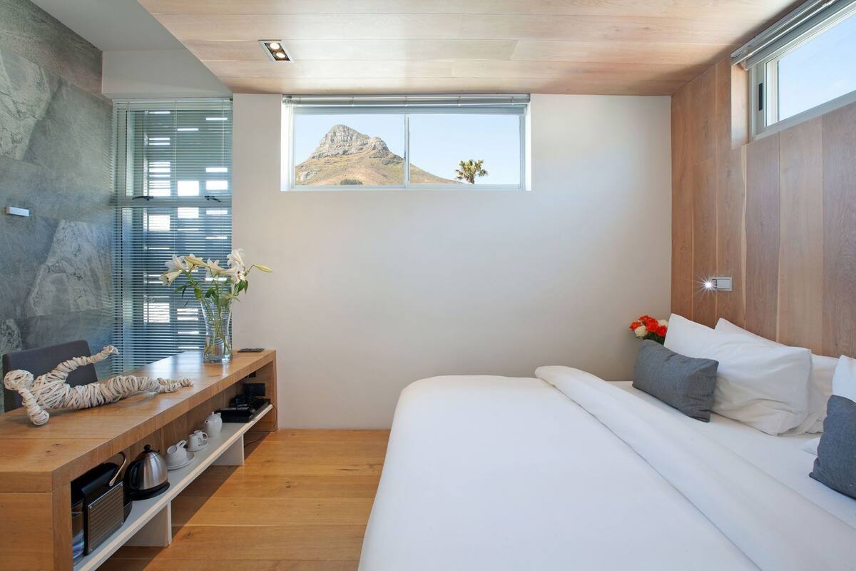A modern bedroom with a wooden headboard, a large window with a mountain view, a bedside table with a vase of flowers, and a bed with white bedding.