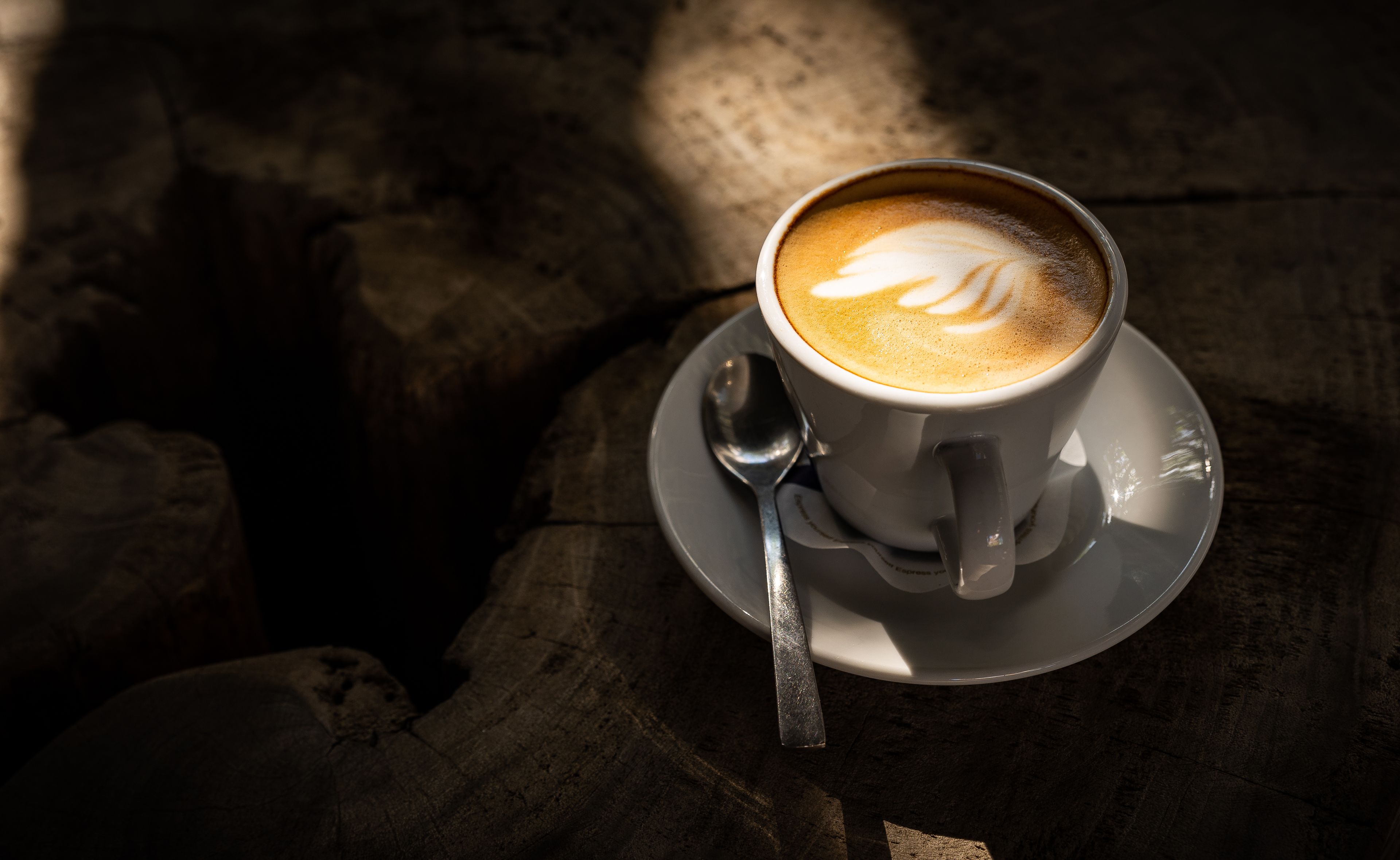 Una taza de café con un diseño de leche, sobre un plato, sobre una superficie de madera y una cuchara.