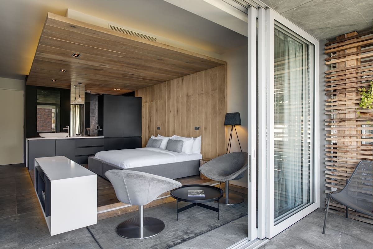 A modern bedroom with a large bed, a bedside table, a chair, and a small table. There is a window with blinds and a view of the outside.
