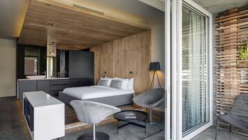 A modern bedroom with a large bed, a bedside table, a chair, and a small table. There is a window with blinds and a view of the outside.
