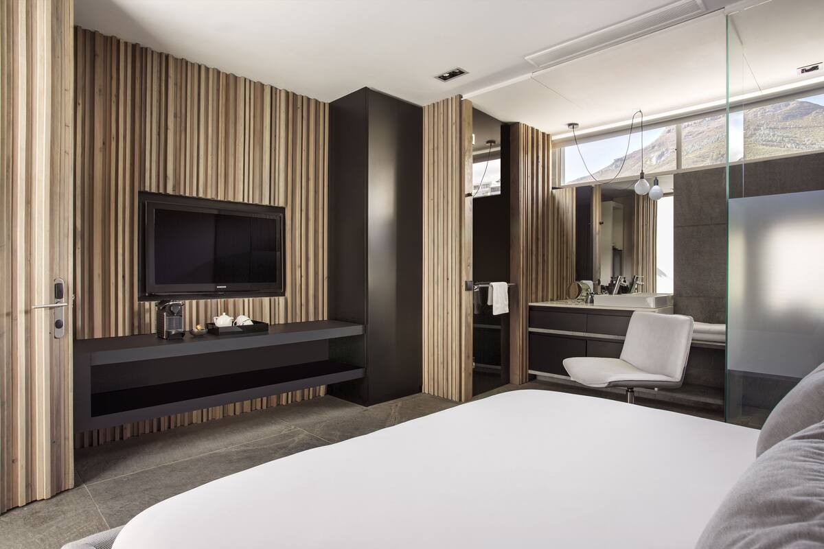 A modern bedroom with a large bed, a TV on a wooden panel wall, a bathroom with a mirror and sink, and a view of mountains through a window.