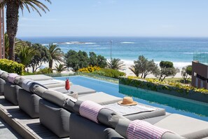 A poolside area with a view of the ocean, a lounge with cushions, and a beach in the background. - POD Camps Bay (Cape Town)